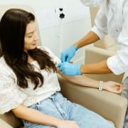 A woman sits in a beige chair while a healthcare worker wearing blue gloves prepares to administer an injection at Infusions By Boss, specialists in IV Infusion Therapy Brisbane, in a professional clinical setting.