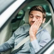 A man in a dress shirt and tie sits in the driver's seat of a car, wearing a seatbelt, eyes closed and rubbing his face, appearing tired or stressed—perhaps in need of an energy boost from Infusions By Boss IV Infusion Therapy Brisbane.