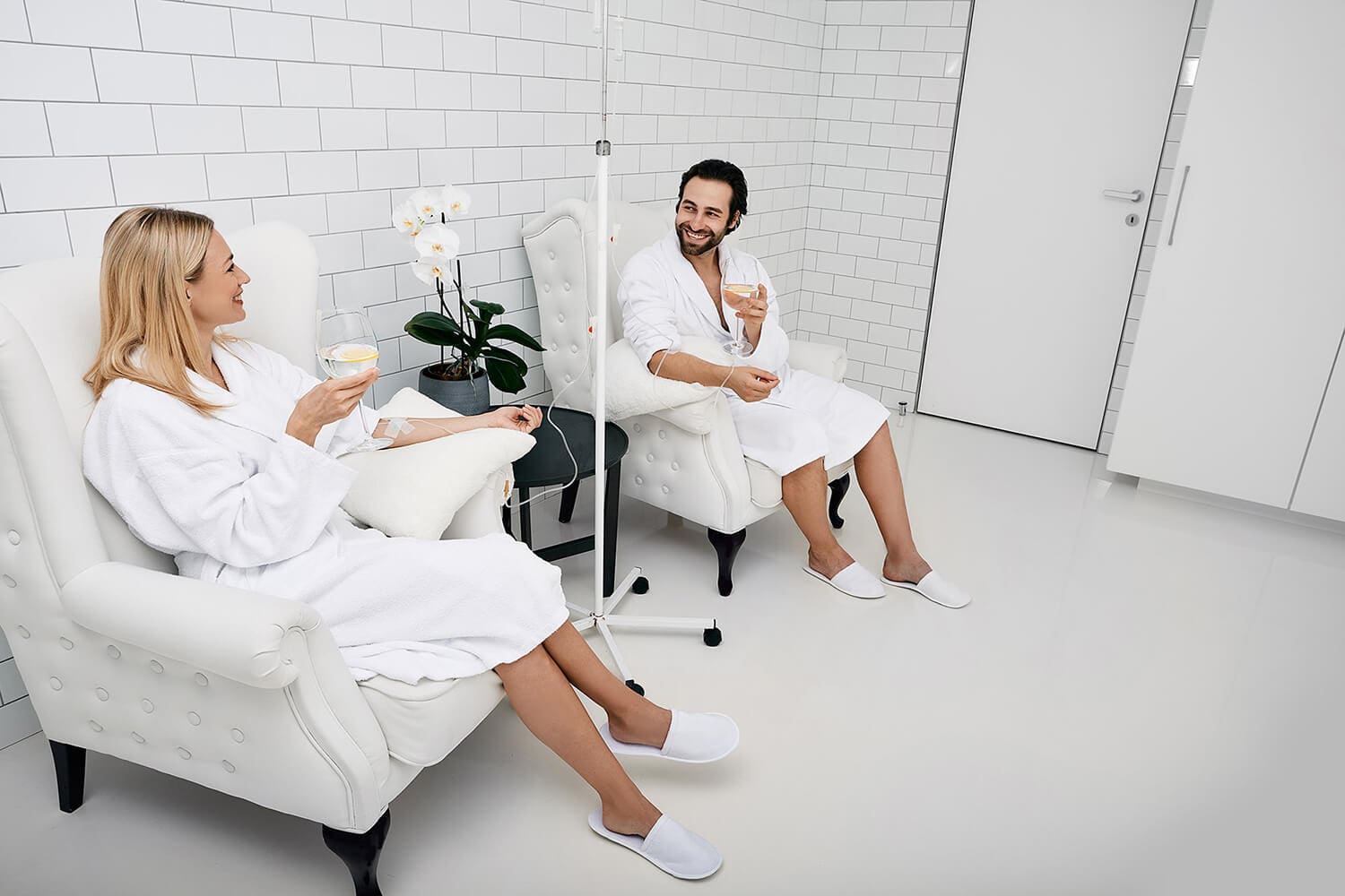 Two people in white robes sit in armchairs in a bright, modern room with white tiled walls at Infusions By Boss. Smiling and holding drinks, one enjoys IV Infusion Therapy Brisbane. A white orchid rests on a small table between them.