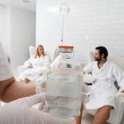 A person wearing gloves holds a large clear bottle of IV fluid, highlighting Infusions By Boss, while a man and woman in white bathrobes chat in armchairs in a bright, tiled room—an inviting scene for IV Infusion Therapy Brisbane.