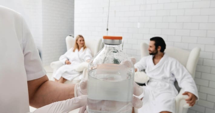A person wearing gloves holds a large clear bottle of IV fluid, highlighting Infusions By Boss, while a man and woman in white bathrobes chat in armchairs in a bright, tiled room—an inviting scene for IV Infusion Therapy Brisbane.