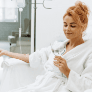 A woman with red hair, wearing a white robe, sits in a clinic chair at Infusions By Boss receiving IV Infusion Therapy Brisbane. She smiles and holds a glass of water with mint, appearing relaxed in a bright, clean medical setting.