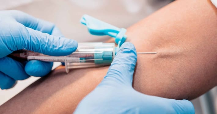 A close-up of a healthcare worker from Boss Infusions wearing blue gloves, inserting a needle into a person's arm to draw blood, with a tourniquet tied above the injection site—showcasing IV Infusion Therapy Brisbane.