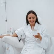 A woman in a white robe sits in a chair at Infusions By Boss, receiving IV Infusion Therapy Brisbane while smiling and holding a glass of wine. The bright, clinical setting suggests a relaxing spa or wellness clinic experience.