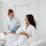 A healthcare professional wearing gloves prepares to draw blood or administer an IV to a smiling woman in a white robe at Infusions By Boss, a leading IV Infusion Therapy Brisbane clinic. She sits comfortably, holding a glass of water.